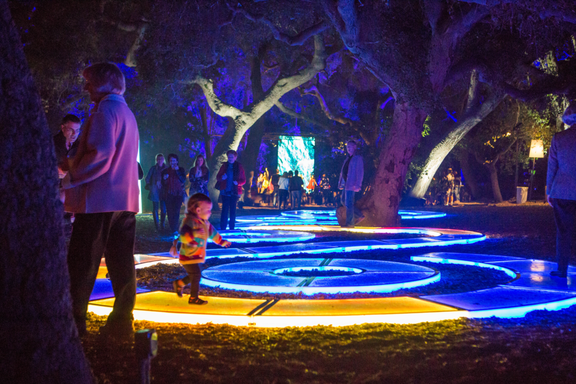 Aqueous (2017, traveling) by Jen Lewin is a large-scale interactive sculpture composed of hundreds of modular LED platforms configured into swirling pathways. By day, the surfaces shift hues and reflect the sky; at night, they respond to touch and motion with glowing, interconnected light trails. Made from code-driven modules derived from the Golden Ratio, each platform senses visitors individually but can link to neighbors to form dynamic visual patterns. Aqueous has toured globally—from Burning Man to Tokyo’s Takanawa Gateway Fest, Sydney’s Vivid, Descanso Gardens, and more—offering immersive play, reflection, and light-driven geometry.