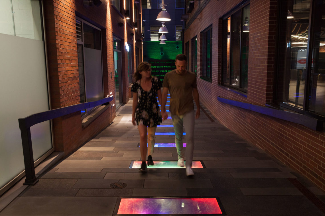 Two people walking across Promenade installation — stepping on glowing circular pads to trigger light