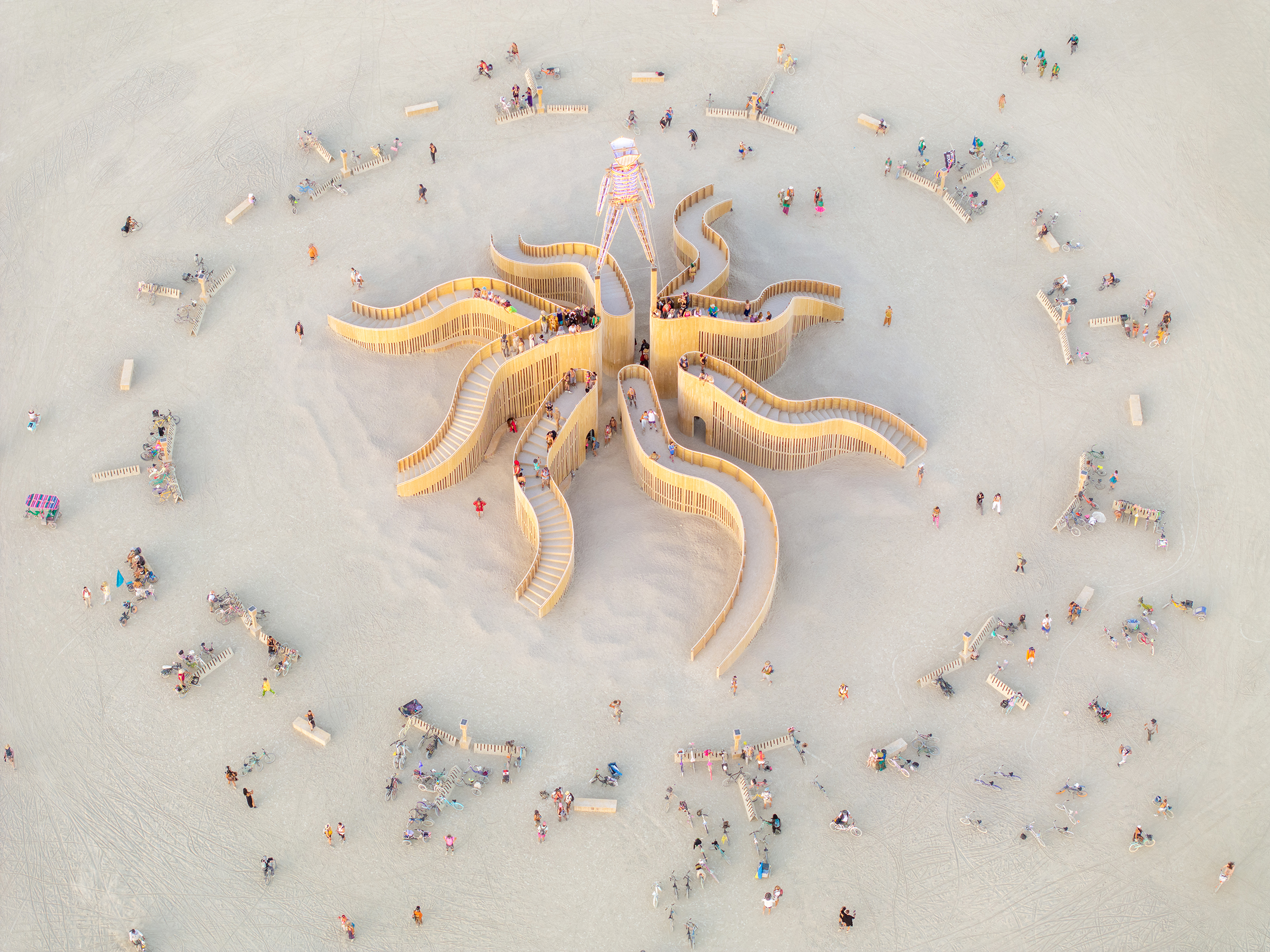 The Other, 2024 Burning Man, Man Pavilion. Photo by Duncan Rawlinson. Photo by Duncan Rawlinson. The Other (2024) by Jen Lewin is the selected Man Pavilion for Burning Man. It comprises eight sweeping, canyon-shaped tentacles, each with its own orientation (inwards, outwards, upwards, away, towards), forming a labyrinth of stairs, ramps, and vantage points. Rather than converge on a single center, the design emphasizes the journey through space and the “in-between”—encouraging meandering, discovery, and multiple perspectives. Beneath the structure lies an open central area with benches and seating, bridging interior and exterior spaces and fostering congregation. The pavilion’s form frames views both outward (toward The Man and The City) and inward (toward shared space), offering a poetic interplay of light, structure, and human participation.